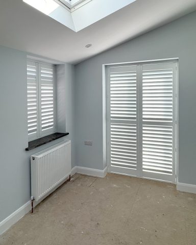 63mm Hardwood shutters in Cardiff. Bright room with light blue walls, wooden shutters, and a radiator beneath a window.