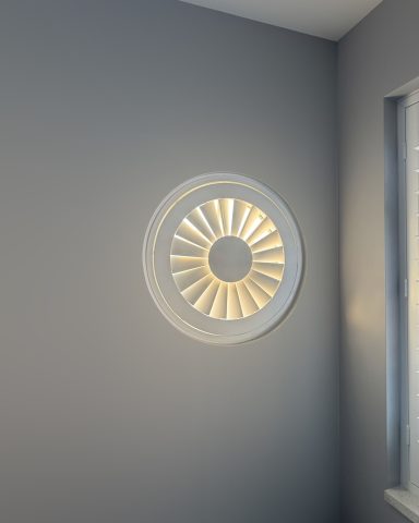 All shapes available. Round window with slatted design, casting light onto a grey wall corner.